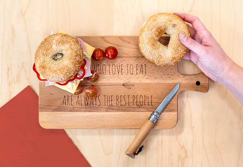 Houten snijplank met handvat, gepersonaliseerd met "WHO LOVE TO EAT ARE ALWAYS THE BEST PEOPLE," getoond met bagels, mes en tomaten.